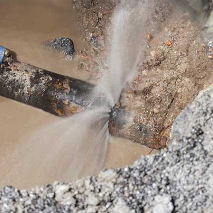 A burst water pipe sprays water into a muddy hole in the ground