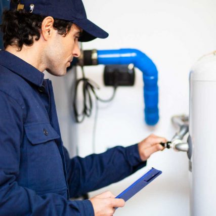 A technician in a blue uniform inspects a large white water heater while holding a clipboard