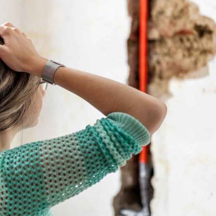 A woman standing, looking at a damaged wall with exposed pipes