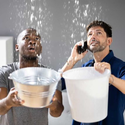 Two men catching water in buckets from a leaking ceiling, with one on the phone